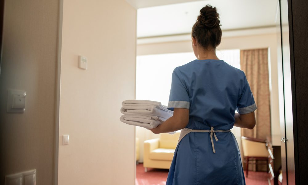 Back view of young brunette chamber maid in uniform carrying stack of clean white towels for guests while entering one of rooms