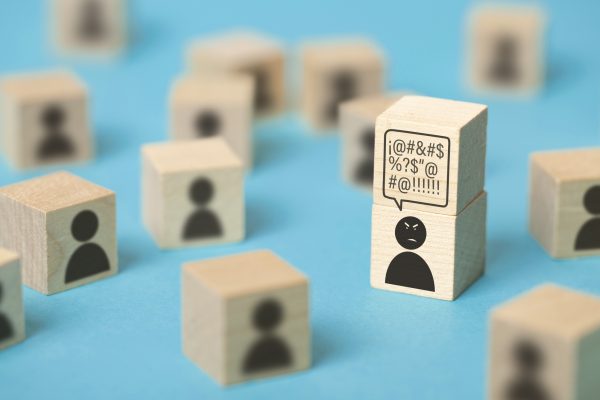 Front view of a group of wooden cubes with icons representing persons. The main focus is on an angry person who has a cube over his head with a thought bubble full of insults