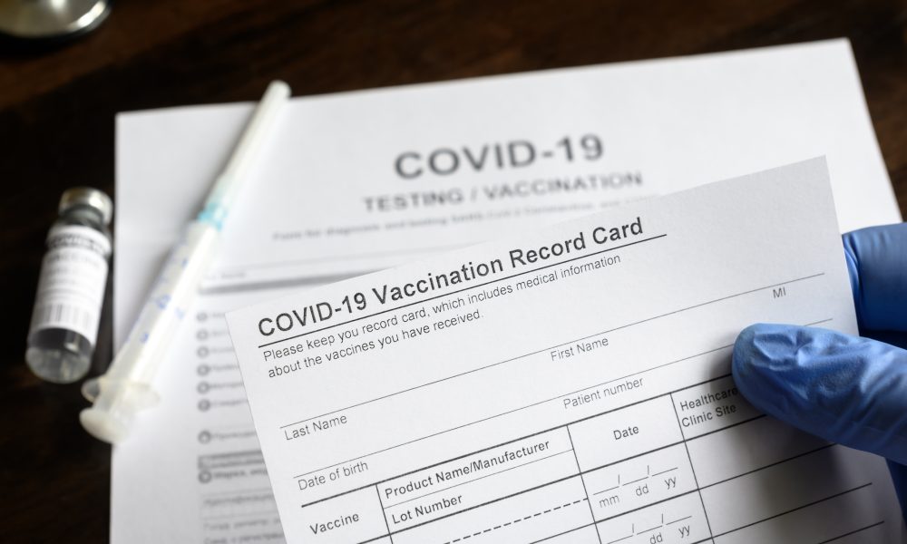 COVID-19 Vaccination Record Card in doctor hand, coronavirus test and vaccine medical form on desk in clinic. Concept of corona virus vaccination certificate, health passport and immunization.