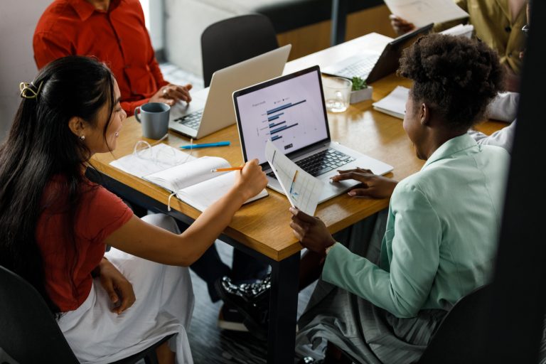 High angle view of two young businesswomen reviewing a document and data on laptop during a meeting with their colleagues.