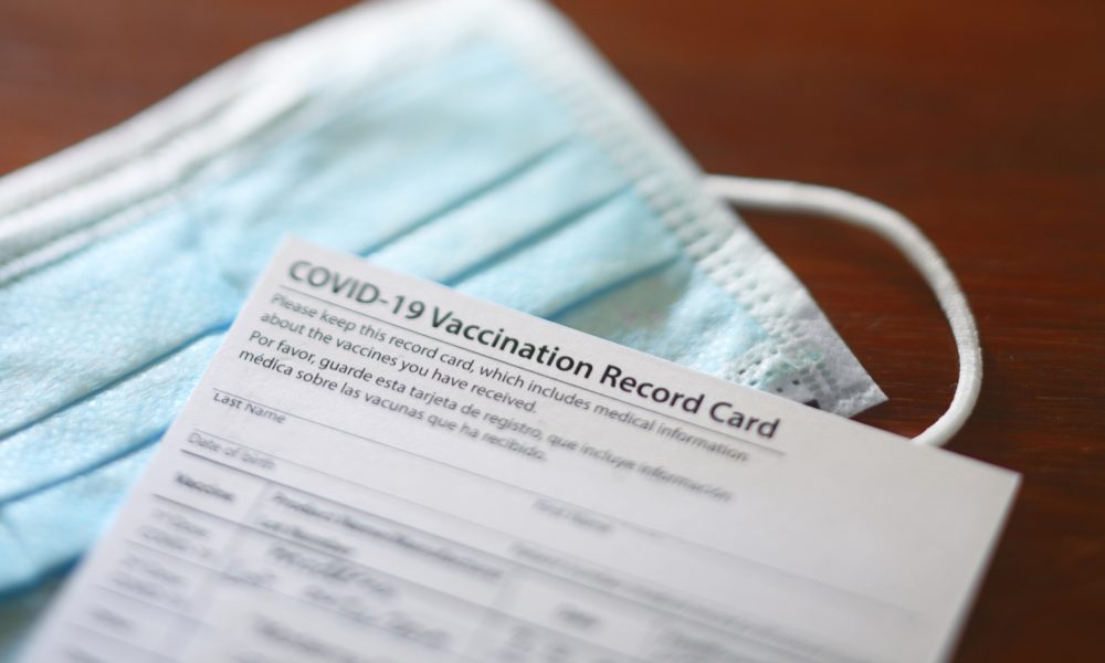 A blank COVID-19 vaccination record card rests on top of a protective face mask.
