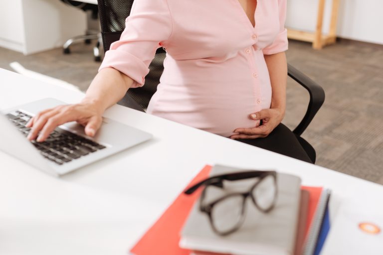 Pleasant pregnant woman sitting at the table and touching her belly while using her laptop