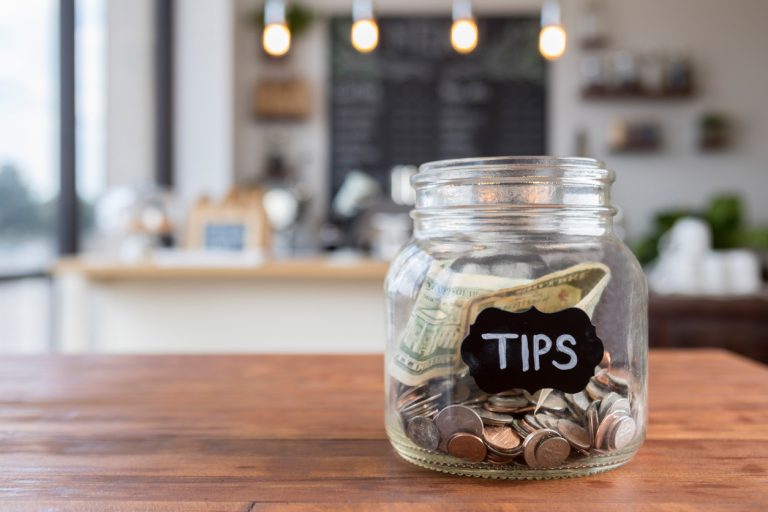 Foreground focus is on a glass jar labeled "tips" in chalk. The jar is sitting to one side on a rustic wooden table, full of coins and bills, with coffee shop scenery in the background.