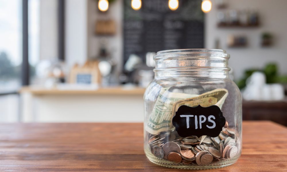 Foreground focus is on a glass jar labeled "tips" in chalk. The jar is sitting to one side on a rustic wooden table, full of coins and bills, with coffee shop scenery in the background.