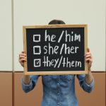 Woman holding chalkboard with list of gender pronouns near color wall
