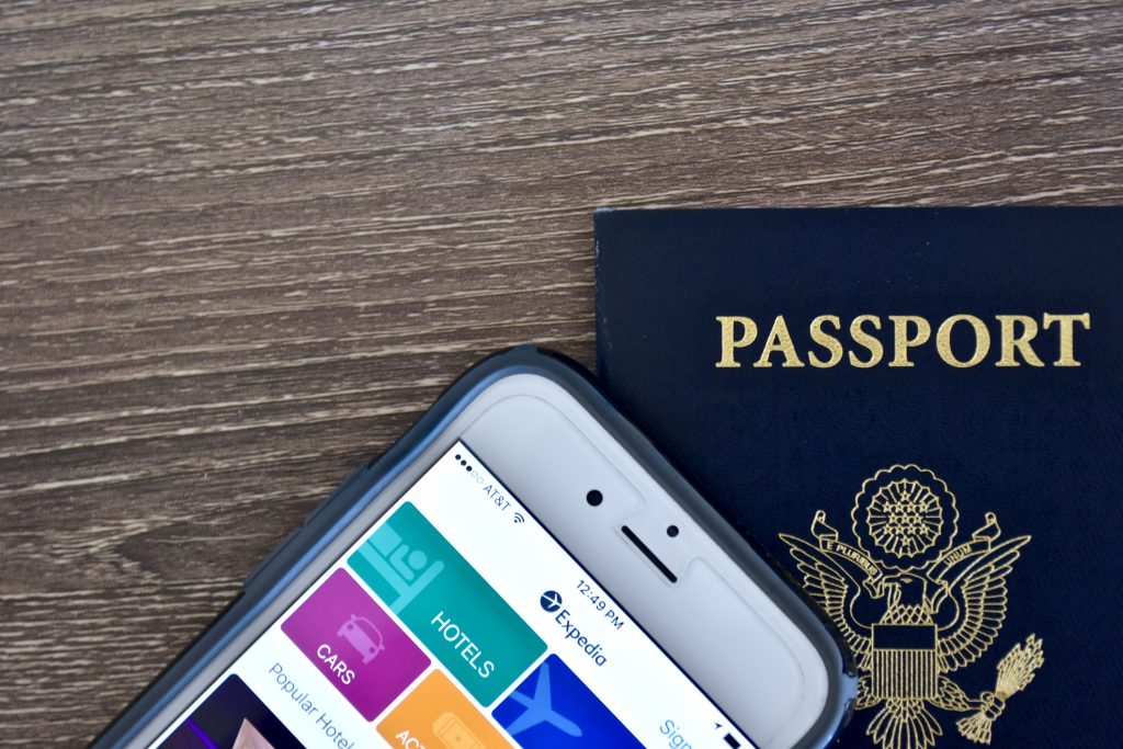 An Apple iPhone 6s displaying the Expedia application homepage on the screen while laying next to an American passport.