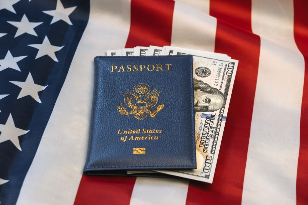 concept for citizenship or travel United States passport holding many US dollar bills and set against Stars and Stripes - HospitalityLawyer.com®