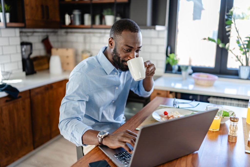 African-American man in a modern kitchen drinking coffee and working on a laptop during breakfast Bright and productive morning scene - HospitalityLawyer.com®