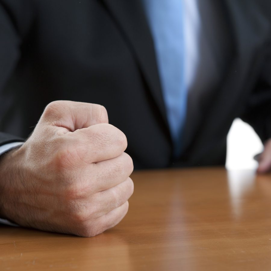 Close-up of a clenched fist resting on a wooden table, with a person in a dark suit and blue tie in the background.