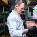 Chef in a white coat with gloves places a tray of raw salmon in a refrigerated storage shelf in a walk-in cooler.