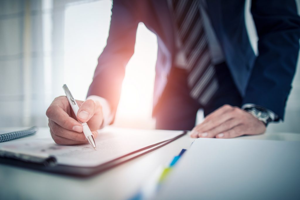 Close-up of a person in a suit signing a document with a white pen at a desk in an office setting