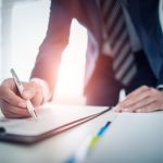Business man signing contract making a deal - HospitalityLawyer.com® Close-up of a person in a suit signing a document with a white pen at a desk in an office setting