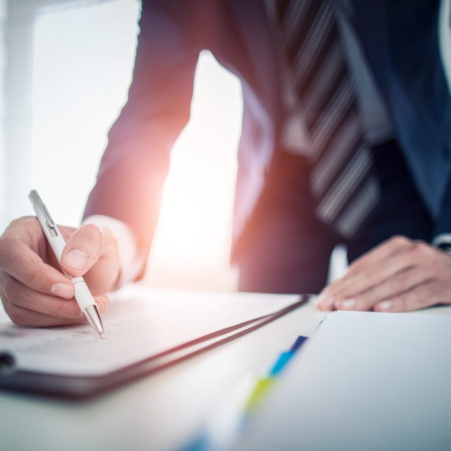 Close-up of a person in a suit signing a document with a white pen at a desk in an office setting