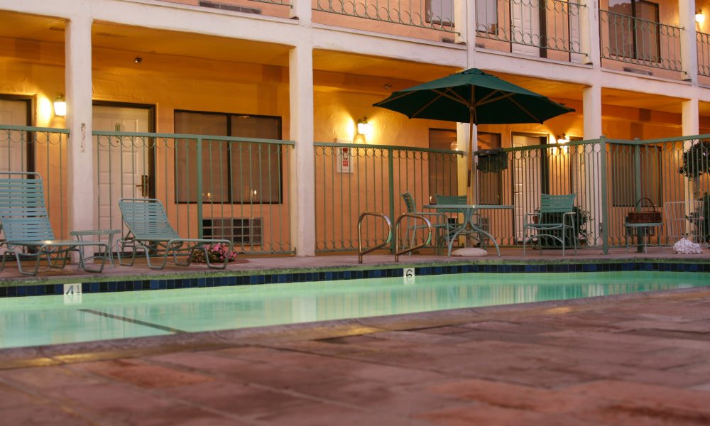 Outdoor motel/pool area with teal lounge chairs, a large green umbrella, and warm wall lights along a two-story building with balconies at dusk