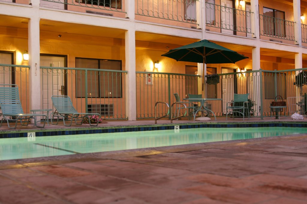 Pool and Rooms at Dusk - HospitalityLawyer.com® Outdoor motel/pool area with teal lounge chairs, a large green umbrella, and warm wall lights along a two-story building with balconies at dusk