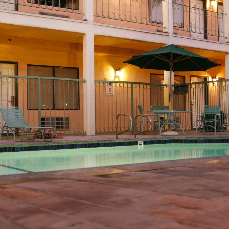 Outdoor motel/pool area with teal lounge chairs, a large green umbrella, and warm wall lights along a two-story building with balconies at dusk